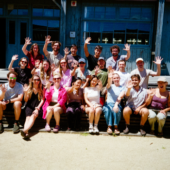 A large group of people sitting and standing outdoors in front of a blue wooden building, smiling and waving at the camera on a sunny day.